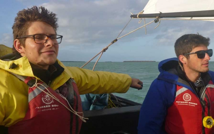 Two people wearing life jackets sit in a sailboat on calm water.